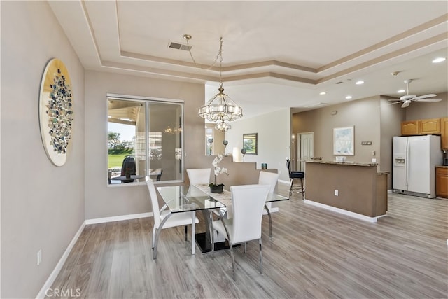 dining space with ceiling fan with notable chandelier, light hardwood / wood-style flooring, and a tray ceiling