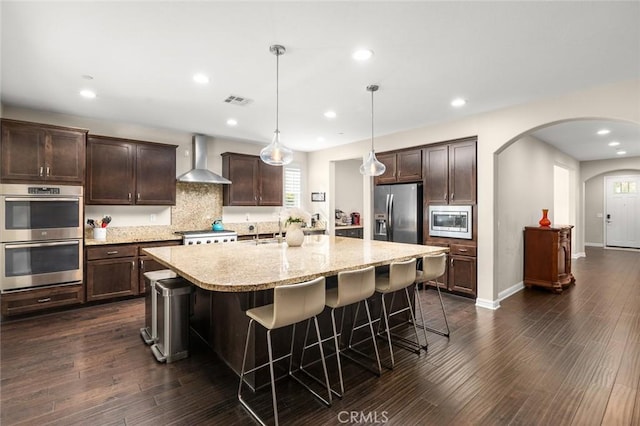 kitchen with dark brown cabinetry, decorative light fixtures, a center island with sink, stainless steel appliances, and wall chimney range hood