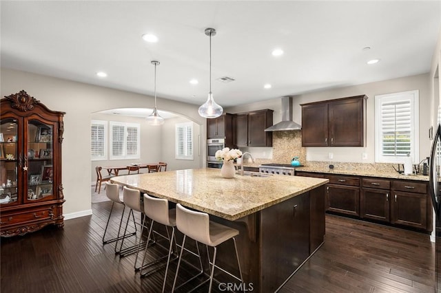 kitchen with wall chimney range hood, sink, hanging light fixtures, light stone counters, and an island with sink