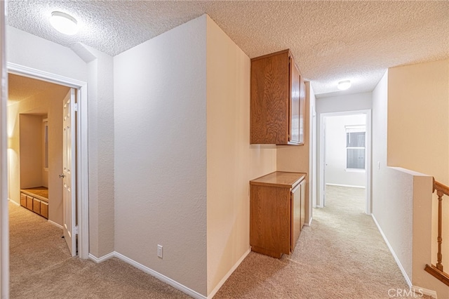 hall with baseboards, a textured wall, a textured ceiling, and light colored carpet