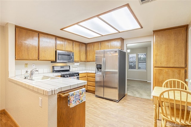 kitchen with stainless steel appliances, tile counters, brown cabinetry, a sink, and a peninsula