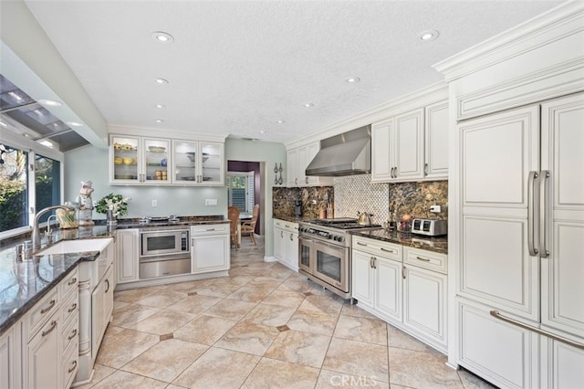 kitchen featuring dark stone countertops, built in appliances, decorative backsplash, and wall chimney range hood