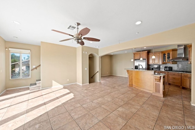 kitchen with wall chimney range hood, light tile patterned floors, stainless steel fridge, a breakfast bar area, and an island with sink