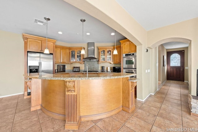 kitchen featuring stainless steel appliances, an island with sink, wall chimney range hood, and light stone counters