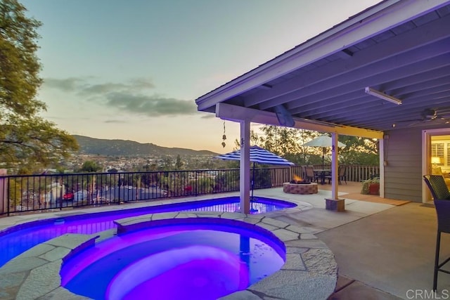 pool at dusk featuring a mountain view, an outdoor fire pit, a patio, and an in ground hot tub