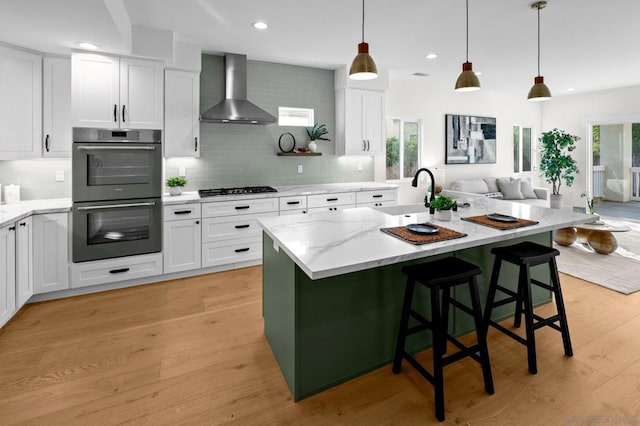 kitchen with white cabinetry, a center island with sink, appliances with stainless steel finishes, pendant lighting, and wall chimney range hood