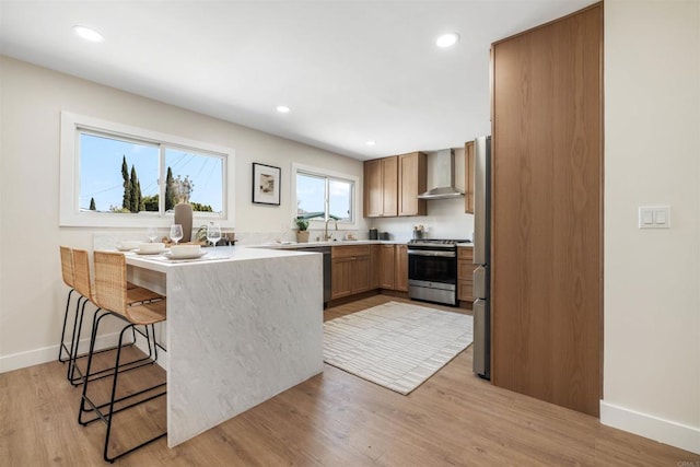 kitchen featuring wall chimney range hood, appliances with stainless steel finishes, a kitchen bar, kitchen peninsula, and light wood-type flooring