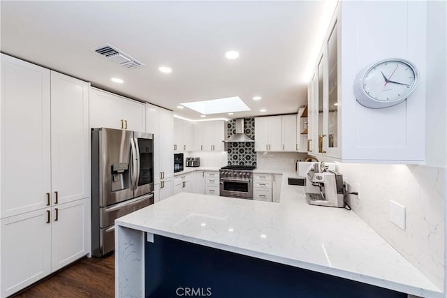 kitchen with appliances with stainless steel finishes, a skylight, white cabinetry, kitchen peninsula, and wall chimney range hood