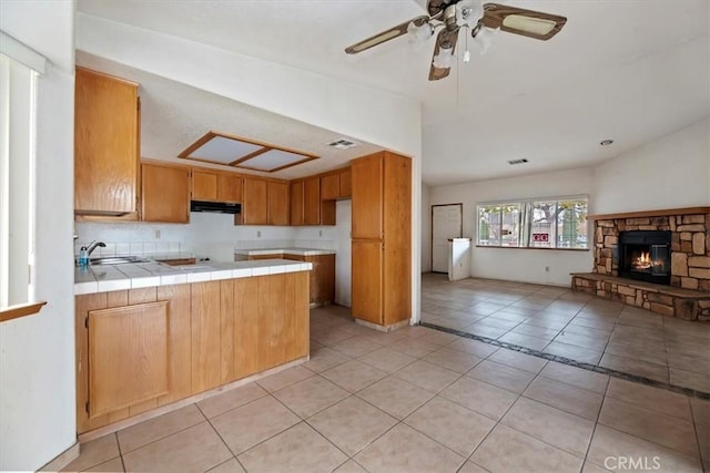 kitchen featuring light tile patterned floors, ceiling fan, a fireplace, tile countertops, and kitchen peninsula