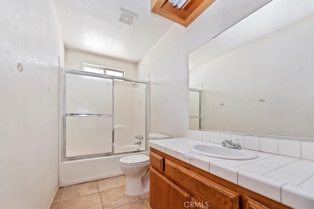 full bathroom with combined bath / shower with glass door, tile patterned flooring, vanity, toilet, and a textured ceiling