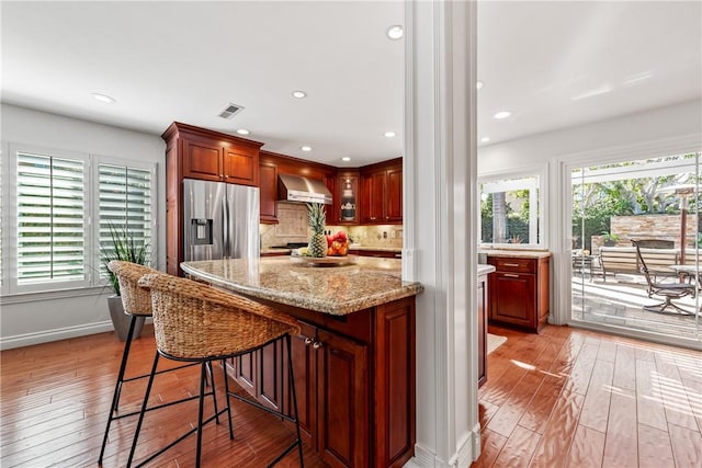 kitchen with stainless steel refrigerator with ice dispenser, wall chimney exhaust hood, backsplash, light wood-type flooring, and a kitchen breakfast bar