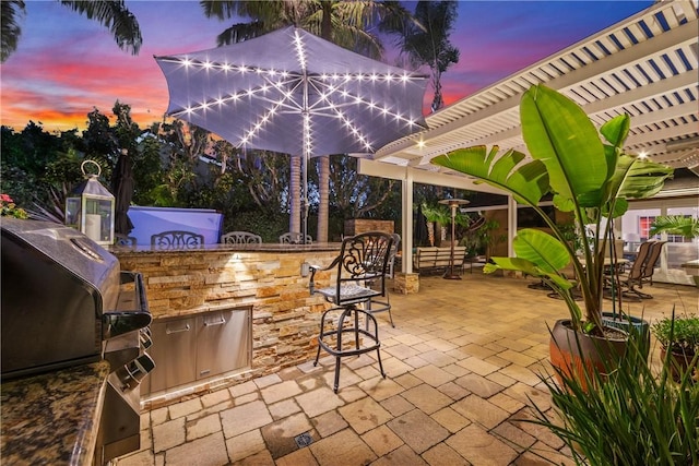 patio terrace at dusk with exterior kitchen, area for grilling, outdoor wet bar, and a pergola