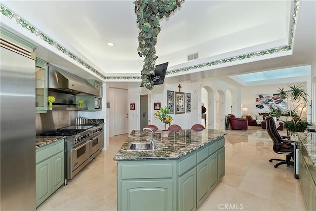 kitchen featuring sink, wall chimney range hood, high end appliances, an island with sink, and green cabinetry