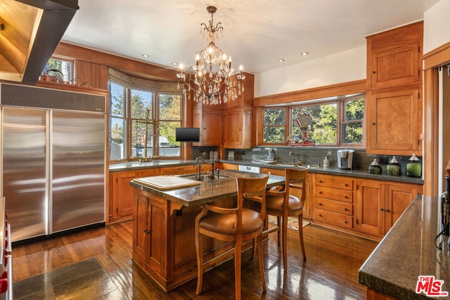 kitchen with a breakfast bar area, built in refrigerator, a kitchen island with sink, backsplash, and decorative light fixtures