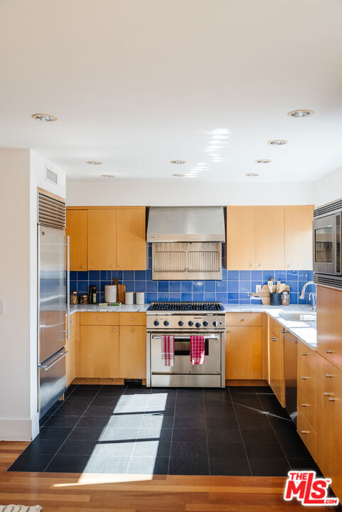 kitchen with wall chimney range hood, sink, built in appliances, and backsplash