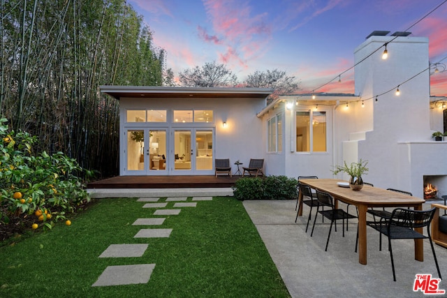 back house at dusk featuring french doors, exterior fireplace, a yard, and a patio