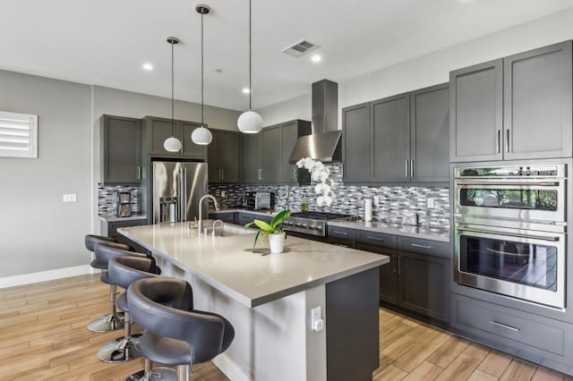 kitchen featuring sink, appliances with stainless steel finishes, a kitchen island with sink, hanging light fixtures, and wall chimney exhaust hood