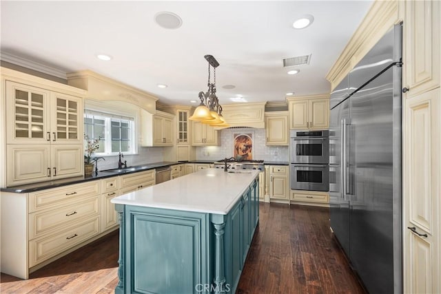 kitchen featuring dark countertops, hanging light fixtures, glass insert cabinets, appliances with stainless steel finishes, and a kitchen island with sink