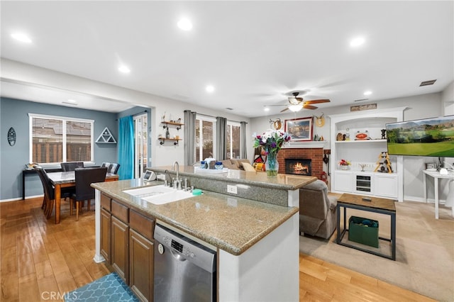 kitchen featuring sink, a brick fireplace, stainless steel dishwasher, an island with sink, and light hardwood / wood-style floors