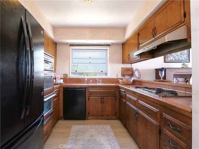 kitchen featuring under cabinet range hood, a sink, light wood-style flooring, black appliances, and brown cabinets