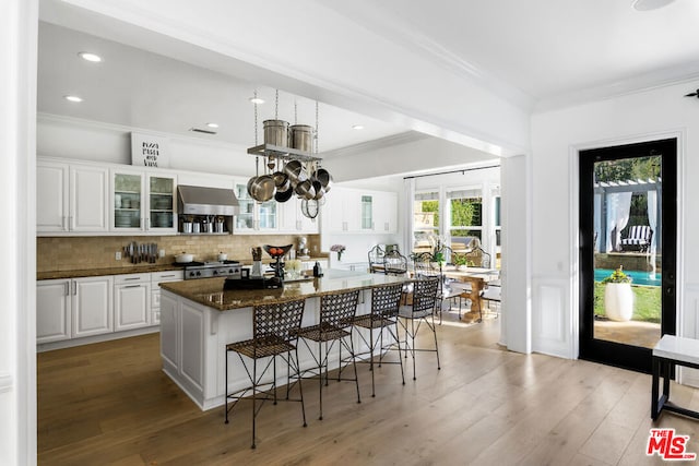 kitchen featuring wall chimney range hood, crown molding, white cabinetry, dark stone countertops, and an island with sink