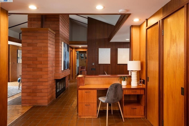 office area with a brick fireplace, vaulted ceiling, wooden walls, and dark tile patterned floors