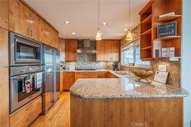 kitchen with brown cabinets, stainless steel appliances, a sink, a peninsula, and wall chimney exhaust hood