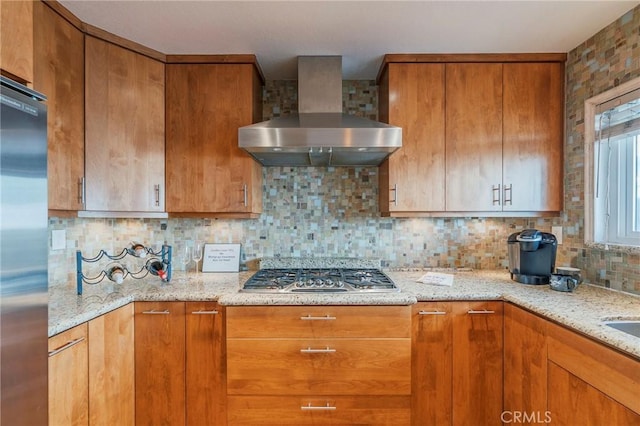 kitchen with appliances with stainless steel finishes, brown cabinets, light stone countertops, wall chimney range hood, and backsplash