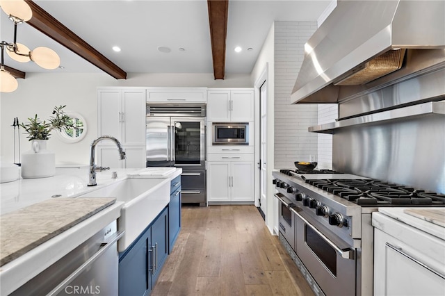 kitchen with decorative light fixtures, white cabinets, built in appliances, blue cabinetry, and wall chimney range hood