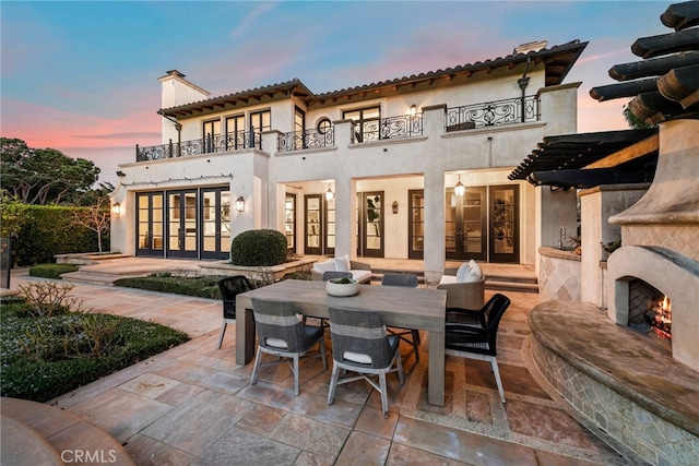 back house at dusk with a patio, a balcony, exterior fireplace, and french doors