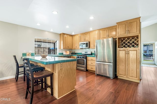 kitchen with dark wood-type flooring, a breakfast bar, appliances with stainless steel finishes, tile counters, and kitchen peninsula