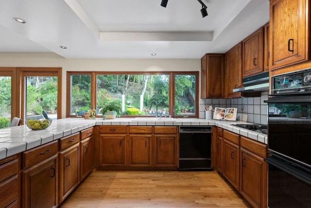 kitchen with sink, light wood-type flooring, a tray ceiling, tile counters, and black appliances