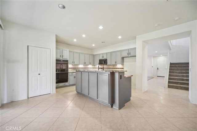 kitchen featuring light tile patterned floors, gray cabinets, double oven, an island with sink, and light stone countertops