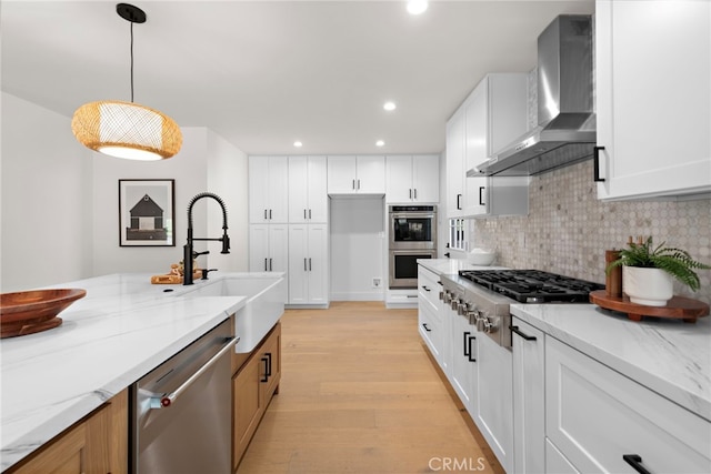 kitchen with wall chimney range hood, hanging light fixtures, stainless steel appliances, light stone counters, and white cabinets