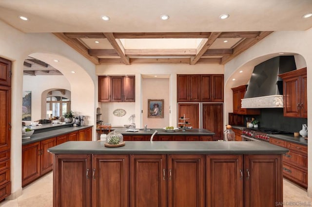 kitchen with a skylight, high end range, coffered ceiling, a center island with sink, and wall chimney exhaust hood