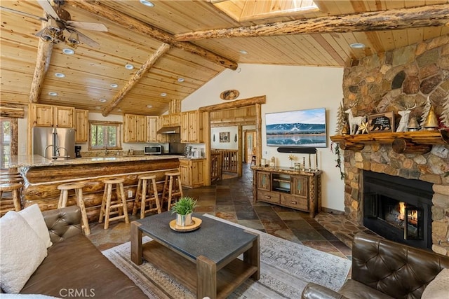 living room with vaulted ceiling with beams, ceiling fan, a stone fireplace, and wooden ceiling