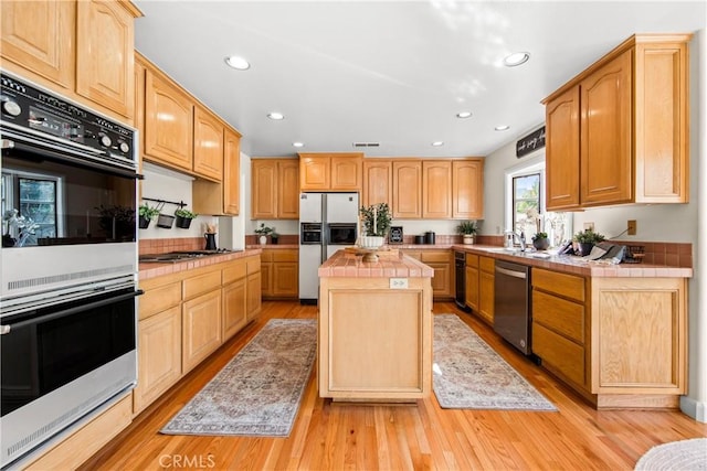 kitchen featuring tile counters, light wood-style flooring, a kitchen island, appliances with stainless steel finishes, and a sink