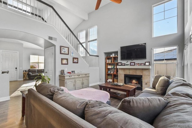 living room featuring ceiling fan, hardwood / wood-style floors, beam ceiling, a towering ceiling, and a tiled fireplace