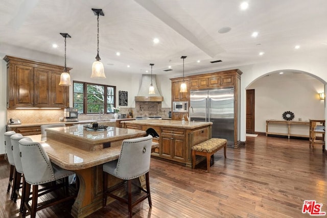 kitchen with a breakfast bar, pendant lighting, a large island with sink, built in appliances, and custom range hood