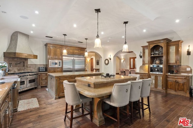 kitchen with a kitchen island, pendant lighting, a breakfast bar area, custom exhaust hood, and built in appliances