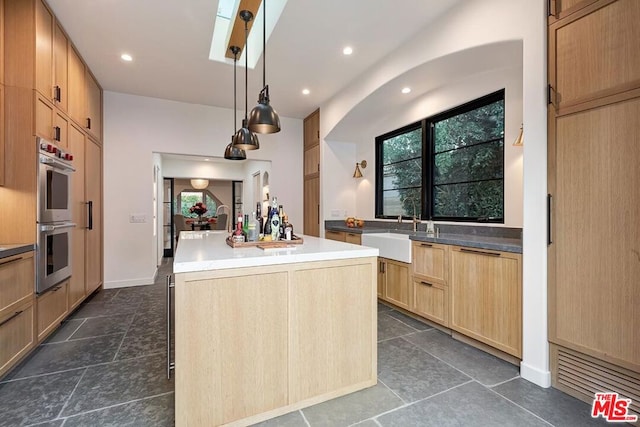 kitchen with light brown cabinetry, an island with sink, sink, hanging light fixtures, and stainless steel double oven