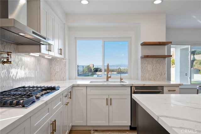 kitchen featuring wall chimney range hood, sink, white cabinetry, light stone counters, and stainless steel gas cooktop