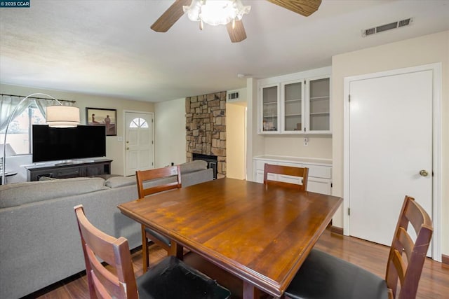 dining room with a stone fireplace, dark wood-type flooring, and ceiling fan