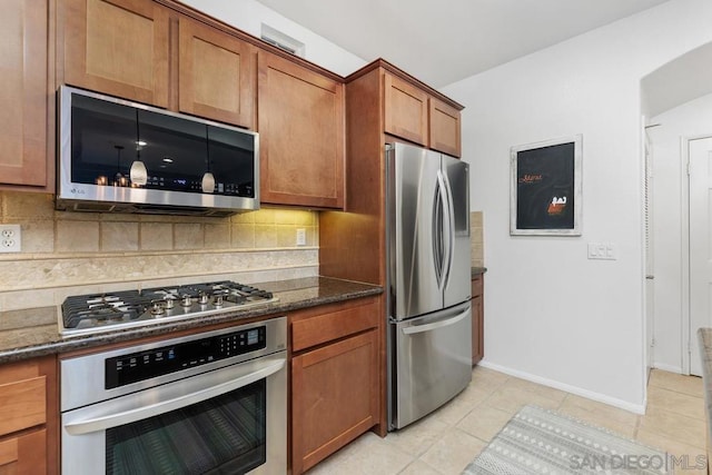 kitchen featuring appliances with stainless steel finishes, light tile patterned floors, decorative backsplash, and dark stone counters