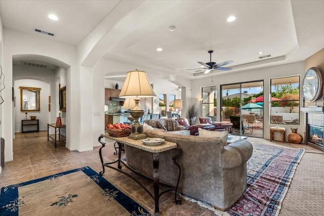 living room featuring a tiled fireplace, ceiling fan, and a tray ceiling