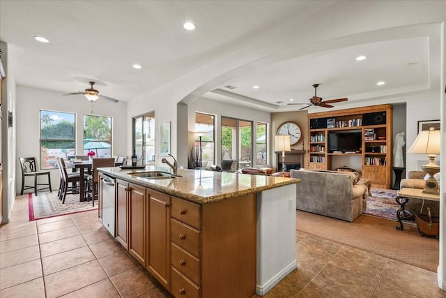 kitchen with sink, a center island with sink, stainless steel dishwasher, ceiling fan, and light stone countertops