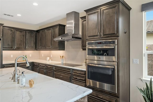 kitchen with wall chimney exhaust hood, light stone counters, tasteful backsplash, double oven, and black electric stovetop