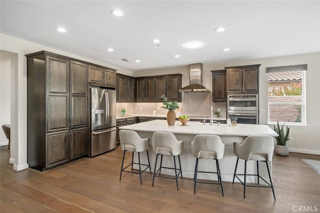 kitchen featuring a center island with sink, a kitchen breakfast bar, wall chimney exhaust hood, and appliances with stainless steel finishes