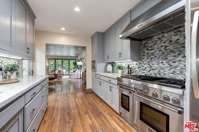 kitchen with wall chimney range hood, gray cabinets, appliances with stainless steel finishes, light stone counters, and light wood-type flooring