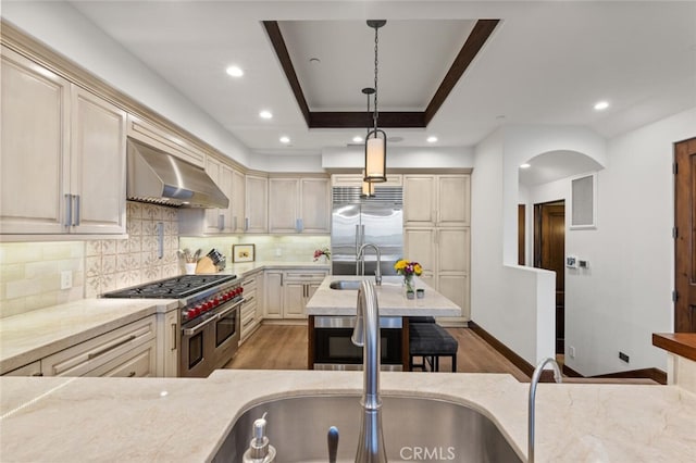 kitchen featuring light stone counters, decorative light fixtures, high quality appliances, cream cabinets, and wall chimney range hood